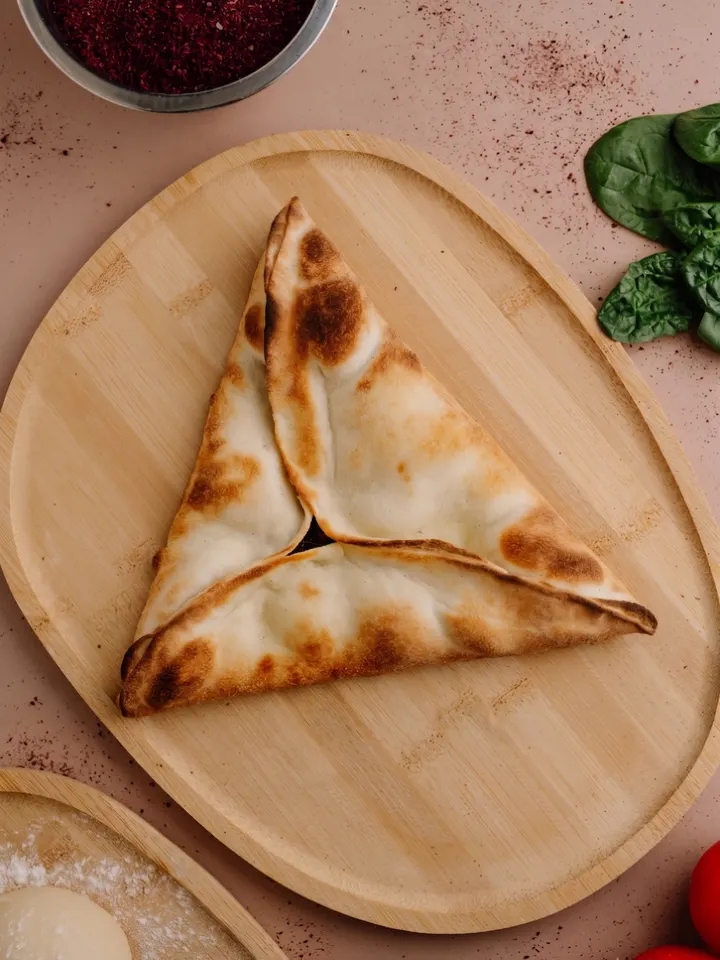 Uno Sbenegh dorato a forma di triangolo su un tagliere di legno, con sommacco e spinaci freschi accanto